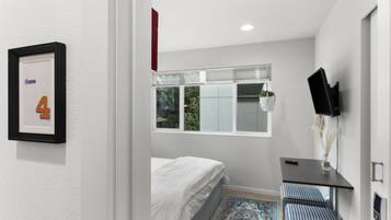 A bedroom with a bed, a desk, a chair, a TV, and a window with blinds.