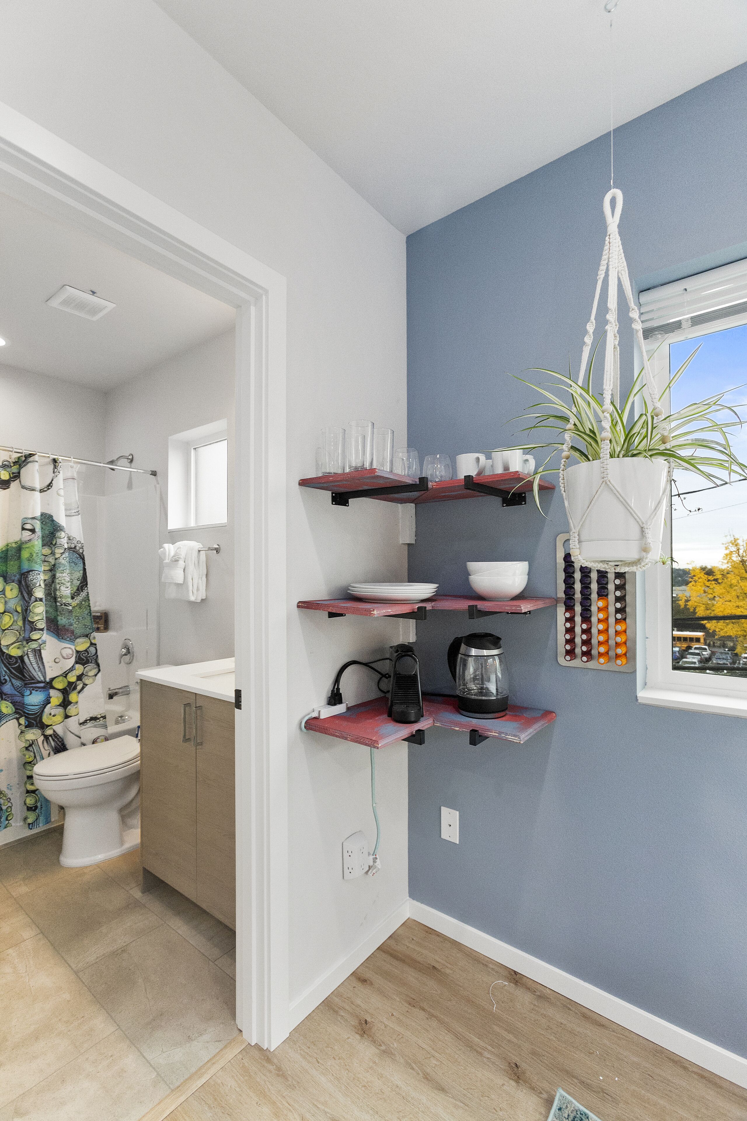 A modern bathroom with a toilet, shower, and a sink. There are shelves with cups and a hanging plant.