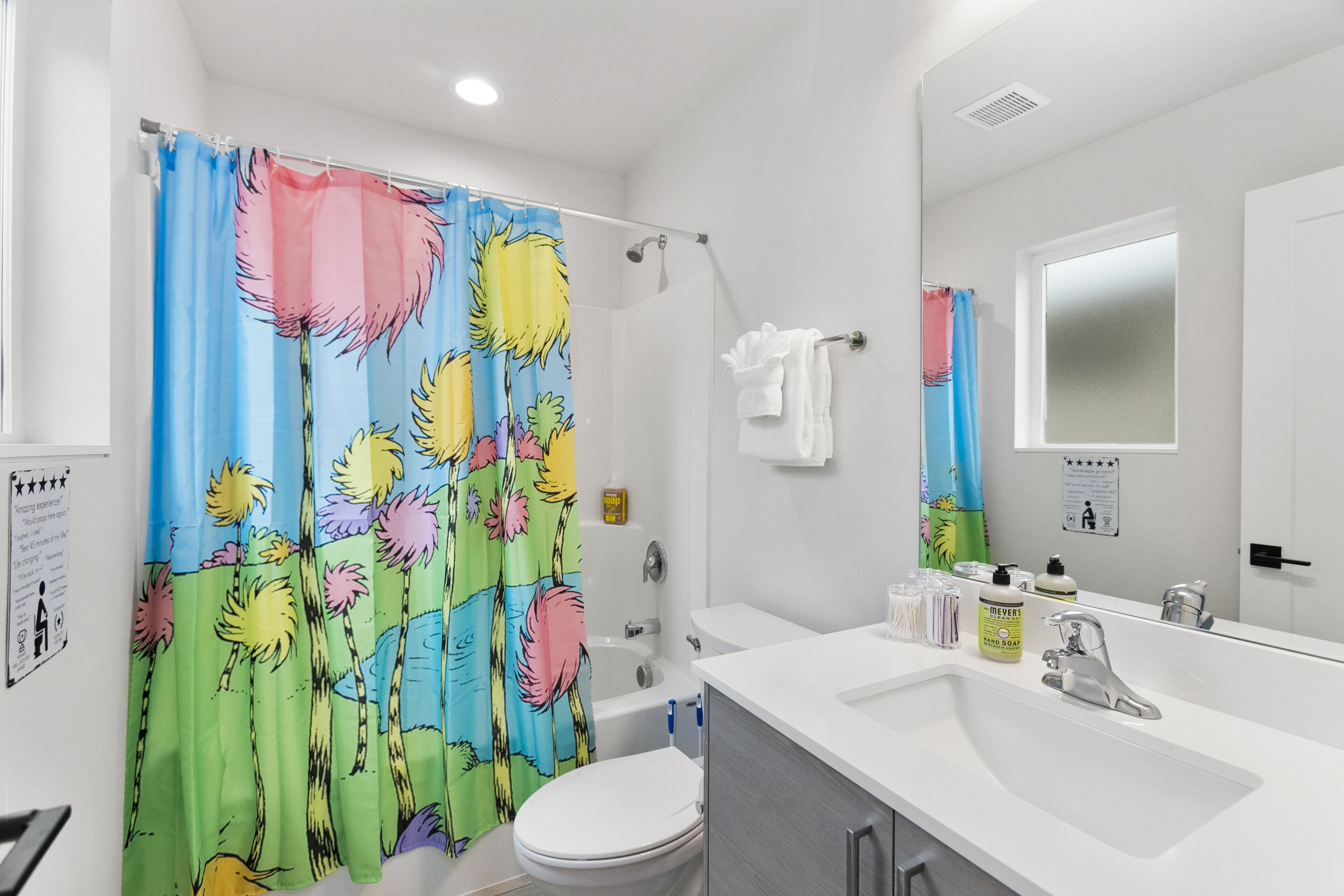 Un baño con cortina de ducha estampada con motivos florales tropicales, un inodoro blanco, una bañera, un lavabo con espejo y un perchero para toallas.