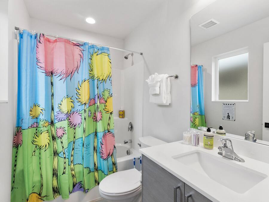 Un baño con cortina de ducha estampada con motivos florales tropicales, un inodoro blanco, una bañera, un lavabo con espejo y un perchero para toallas.