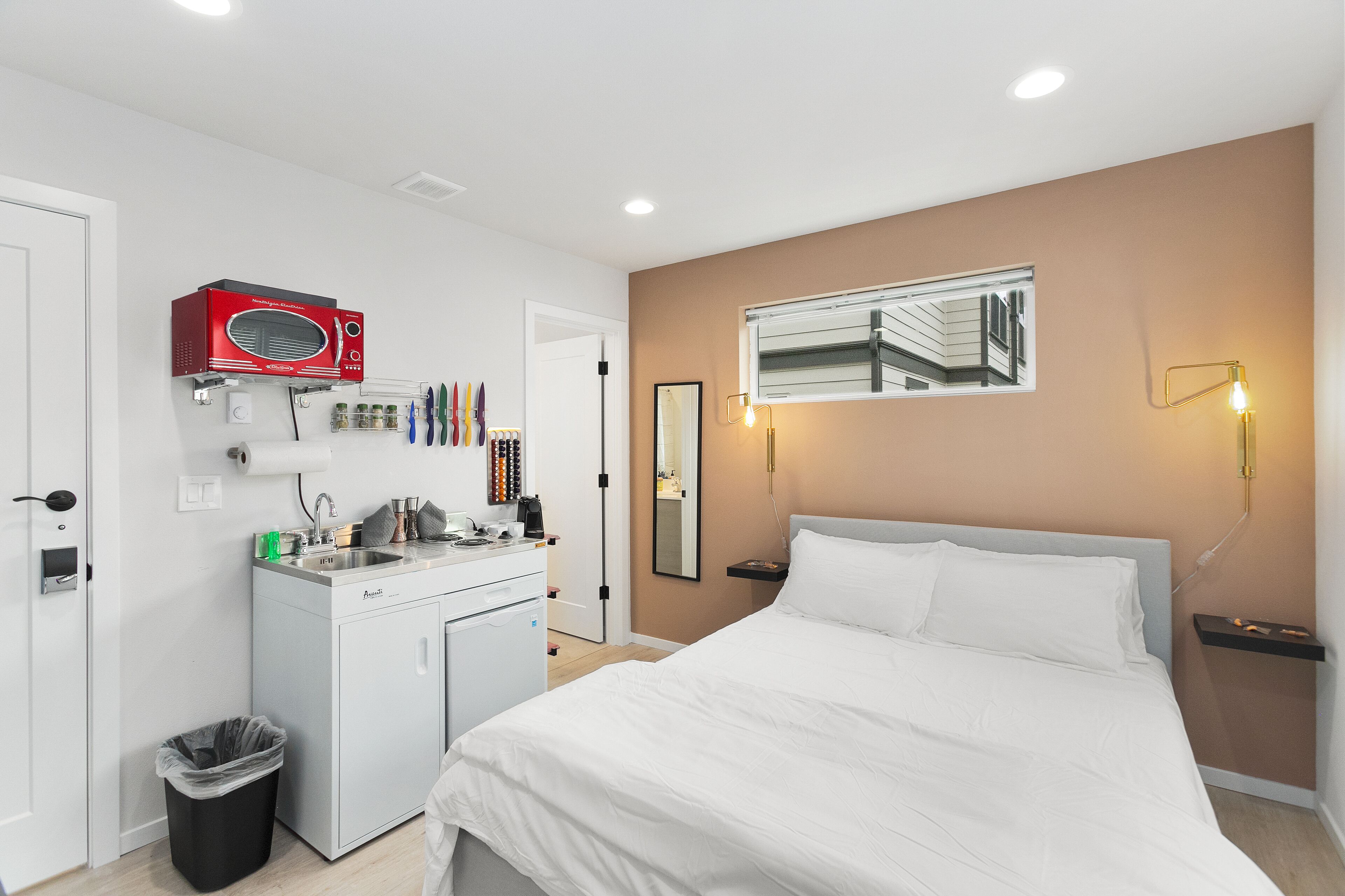 A hotel room with a bed, a small kitchenette, a microwave, a trash bin, and a mirror on the wall.