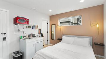 A hotel room with a bed, a small kitchenette, a microwave, a trash bin, and a mirror on the wall.