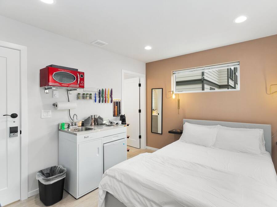 A hotel room with a bed, a small kitchenette, a microwave, a trash bin, and a mirror on the wall.