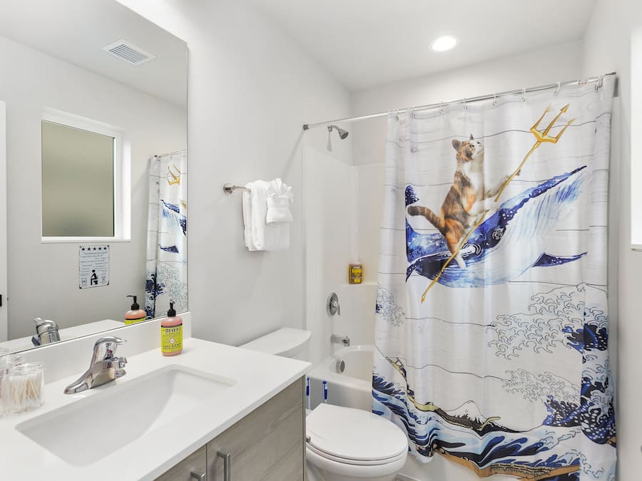 Baño con lavamanos blanco, espejo e inodoro. Hay una cortina de ducha con diseño de olas.