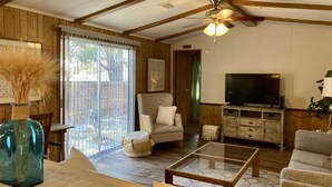 A spacious living room with a high ceiling, wooden beams, a ceiling fan, a large TV, and a glass coffee table. - Convenient NE Albuquerque location, right off I-25!! (Albuquerque)