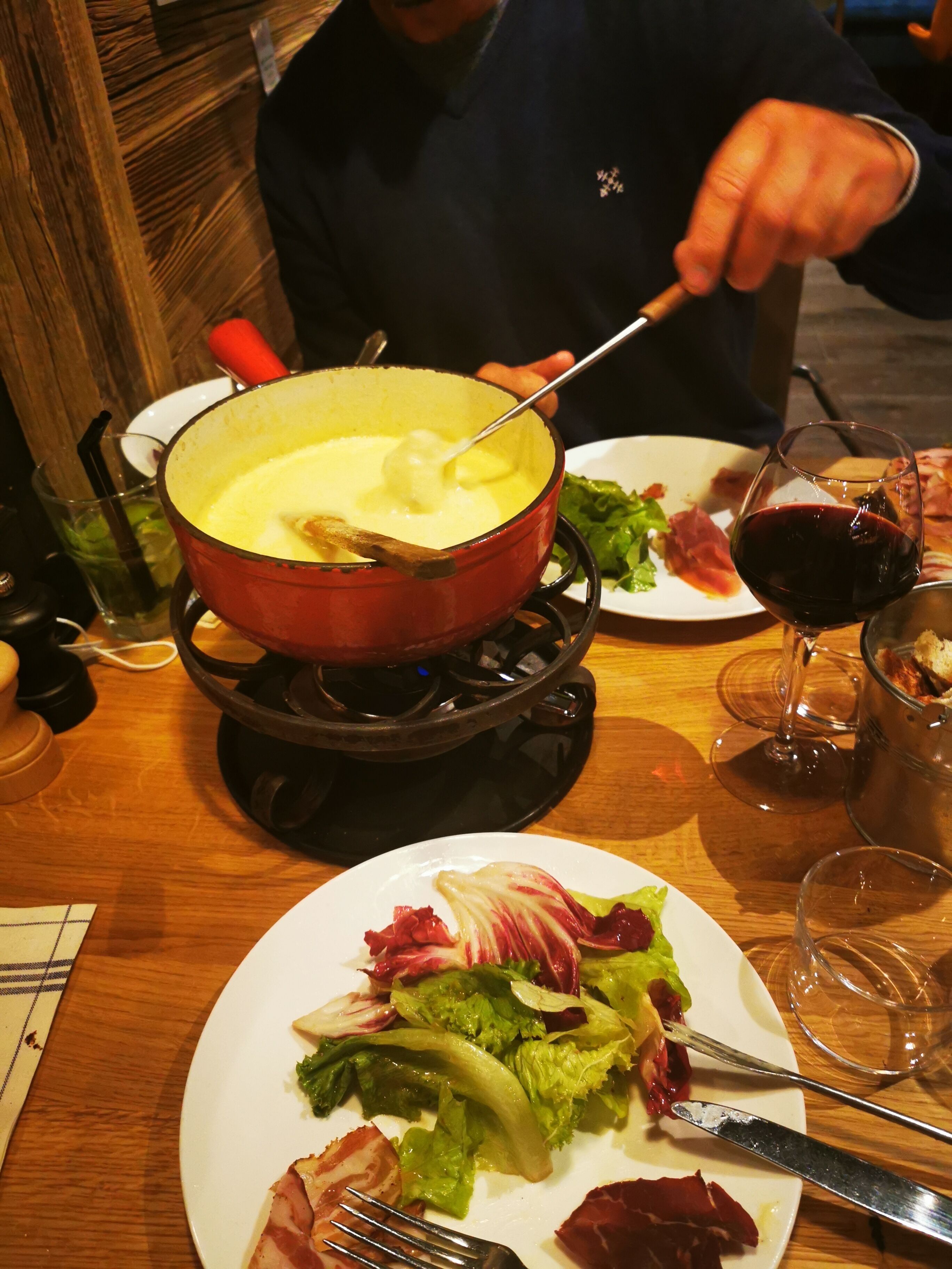 Una mesa de comedor con una olla roja de fondue, un plato de comida y una copa de vino tinto.
