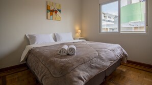 Un lit bien fait, recouvert d’une couverture marron et agrémenté de deux serviettes enroulées, une lampe de chevet et une fenêtre donnant sur des immeubles.