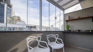 Une pièce moderne avec de grandes fenêtres, deux chaises blanches, une petite étagère ornée d’objets décoratifs et une vue sur le paysage urbain.