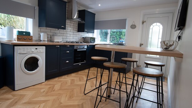 A modern kitchen with a built-in washing machine, dark cabinets, wooden flooring, and bar stools. - Beautiful 200 year old Cottage near St Andrews (Markinch)