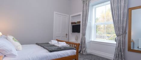 A bedroom with a wooden bed, grey carpet, a white door, a window with grey curtains, and a mirror on the wall.