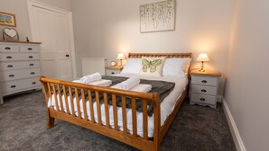 A neatly made bed with a wooden headboard, two bedside tables with lamps, a dresser with drawers, and a framed picture on the wall. - Beautiful 200 year old Cottage near St Andrews (Markinch)