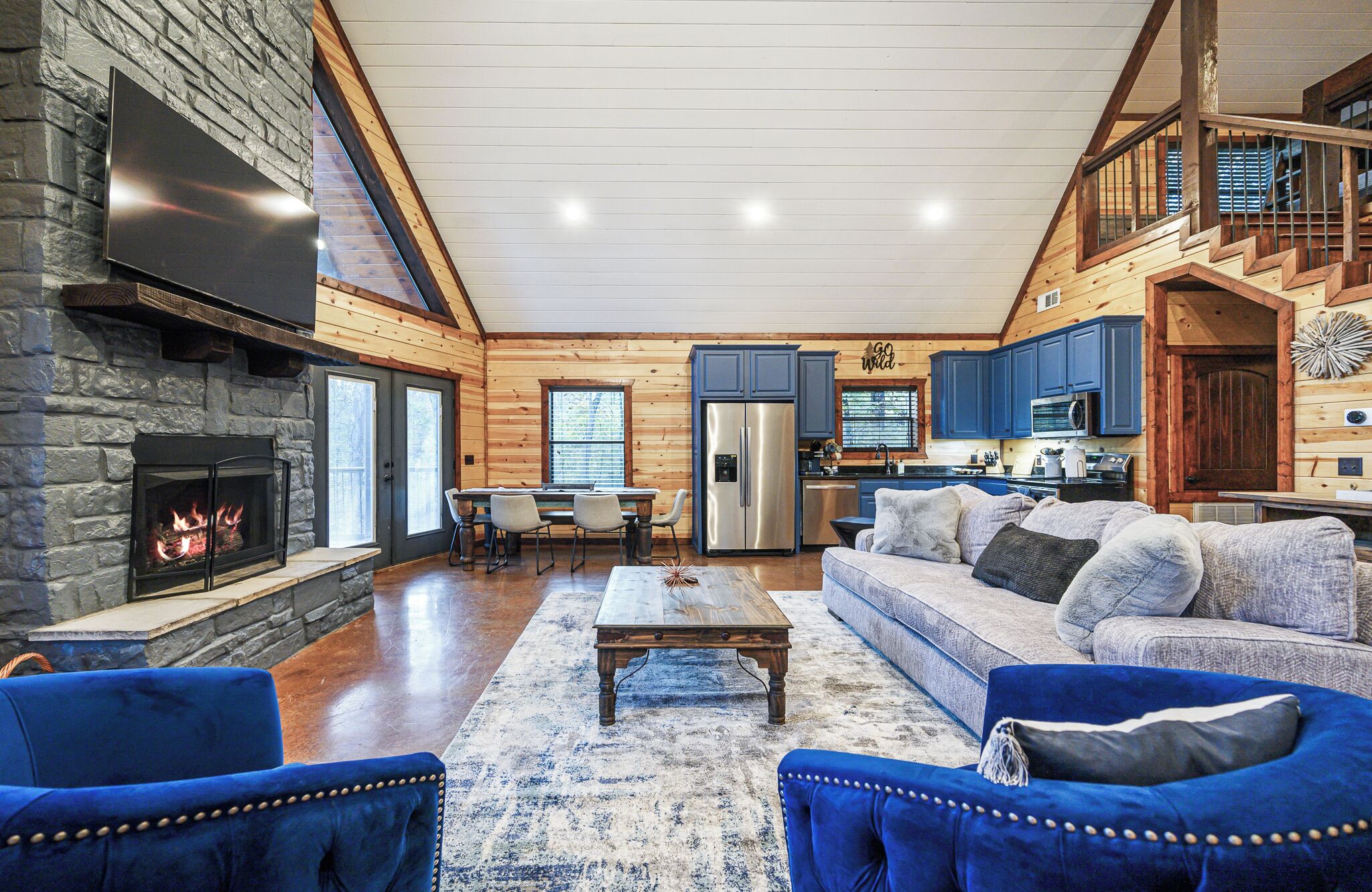 A spacious living room with a stone fireplace, a dining area, a kitchen with blue cabinets, and a staircase leading to an upper level.