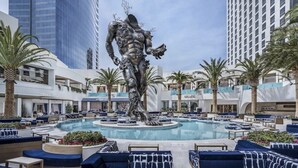 A hotel pool area with a central statue, palm trees, and modern buildings in the background. - Immaculate Views in Las Vegas High Rise Corner Unit  (Las Vegas)