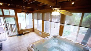 A hot tub in a wooden room with large windows, a wooden deck outside, and a ceiling fan.