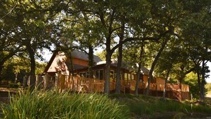 A wooden cabin with a fenced deck, surrounded by trees and a body of water.