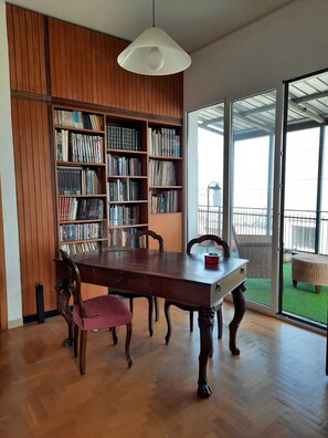 A dining area with a wooden table and chairs, a bookshelf filled with books, and a large window with a view of a green outdoor space.