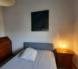 A bedroom with a bed, a nightstand with a lamp, a wooden dresser, and a painting on the wall.