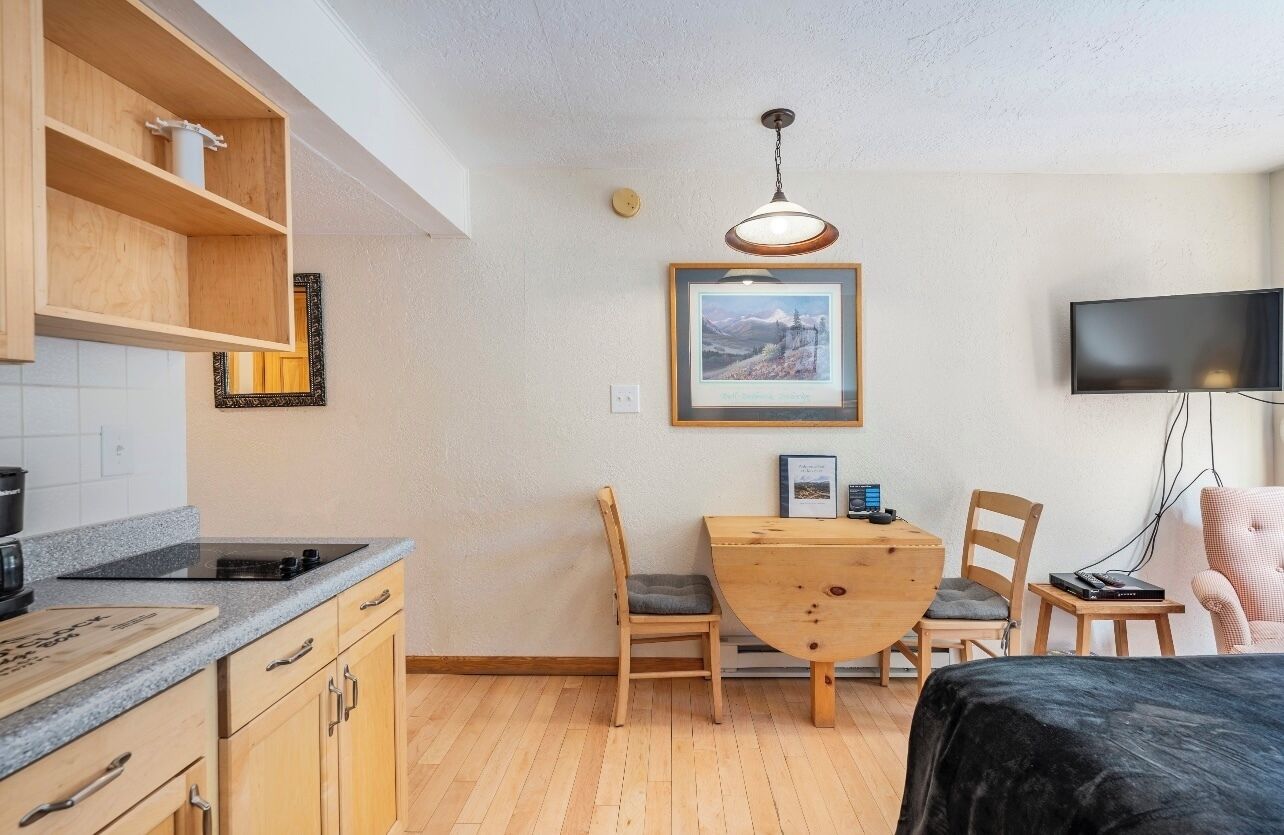 Een compacte kamer met een kitchenette, een eettafel met stoelen, een bed, een televisie en een gekaderd schilderij aan de muur.