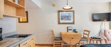 Een compacte kamer met een kitchenette, een eettafel met stoelen, een bed, een televisie en een gekaderd schilderij aan de muur.