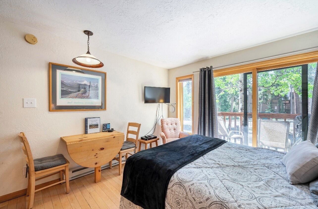 Ein Schlafzimmer mit Bett, Schreibtisch, Stuhl, Fernseher und einem großen Fenster mit Blick auf Bäume.