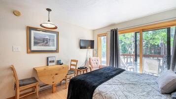 Une chambre à coucher comprenant un lit, un bureau, une chaise, un téléviseur et une grande fenêtre donnant sur la forêt.