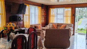 A cozy living room with a sofa, chairs, a dining table with a vase of sunflowers, and a television on the wall. - Private Valley View Cabin near Irazú - 100 Mbps WiFi & Full Kitchen (San Juan de Chicuá)