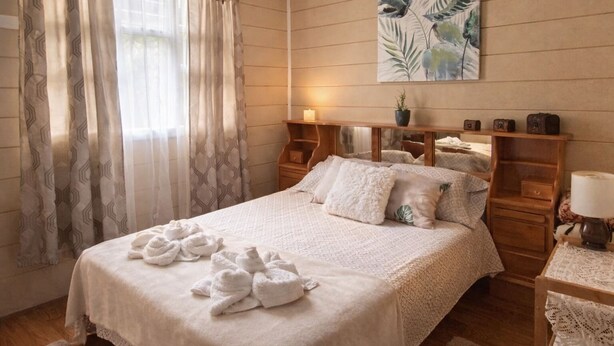 A neatly made bed with towels, a wooden nightstand with a lamp, and a wall-mounted shelf with decorative items. - Private Valley View Cabin near Irazú - 100 Mbps WiFi & Full Kitchen (San Juan de Chicuá)