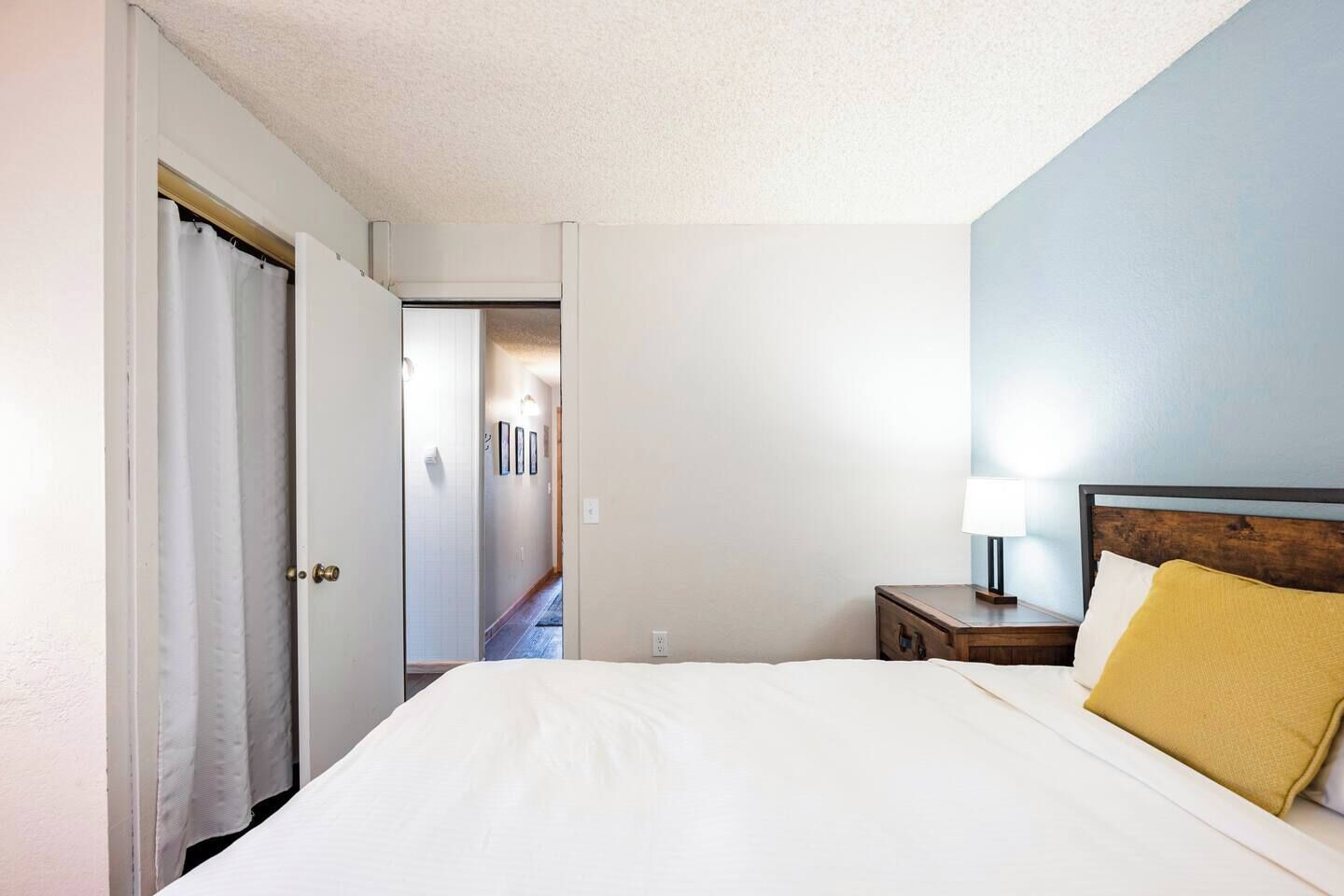 Een hotelkamer met een bed, een nachtkastje met een lamp en een deur die naar een gang leidt.