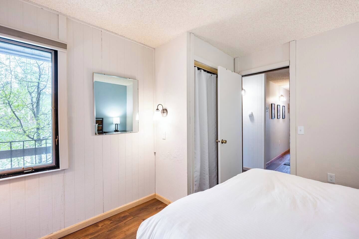 A bedroom with a large window, a mirror, a door leading to a hallway, and a bed.
