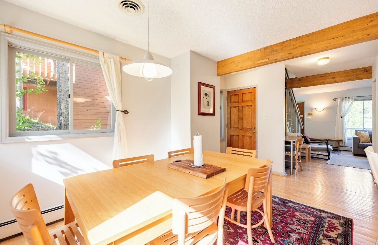 A dining area with a wooden table and chairs, a hanging light fixture, and a view of a garden through a window.