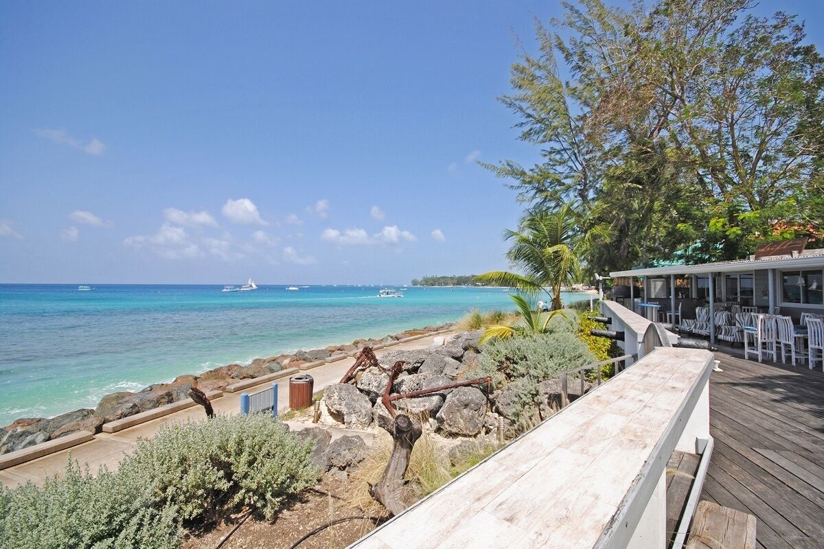A wooden deck overlooking a beach with clear turquoise waters, a rocky shoreline, and a restaurant with outdoor seating.