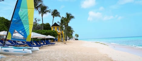 A beach with lounge chairs, a sailboat, and a yellow building.