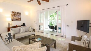 Un salon comprenant un canapé, une table basse et un téléviseur. On aperçoit également un coin repas avec une table et des chaises, visibles à travers une porte vitrée.