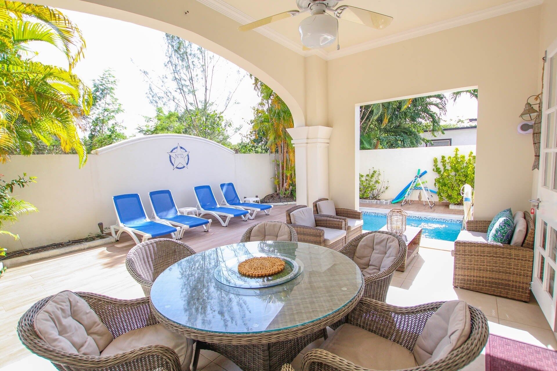 A sunlit patio with a pool, wicker furniture, and blue lounge chairs.