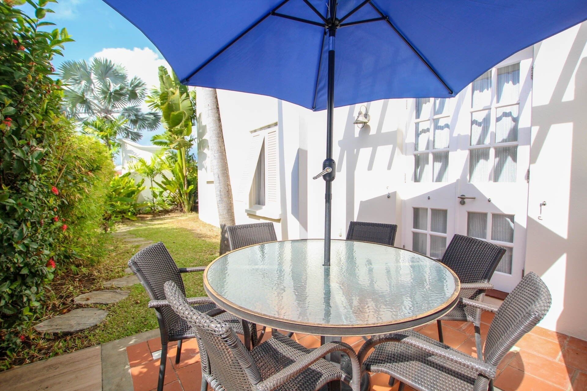 A round glass table with wicker chairs under a blue umbrella, surrounded by a white building with windows and a garden with tropical plants.