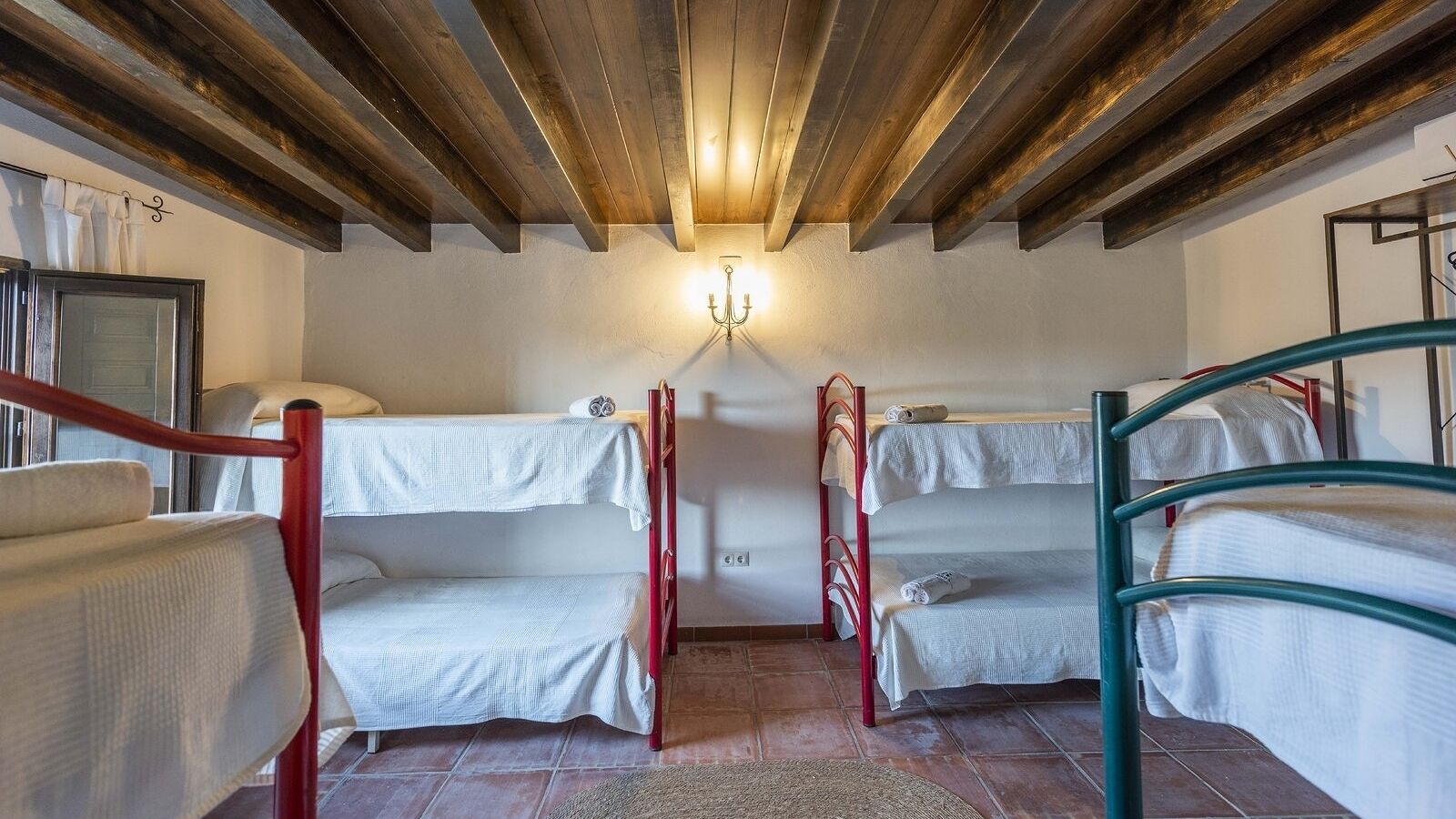 Ein Schlafsaalzimmer mit Etagenbetten, einer Holzbalkendecke, einer Wandleuchte und einem Fenster mit Vorhängen.