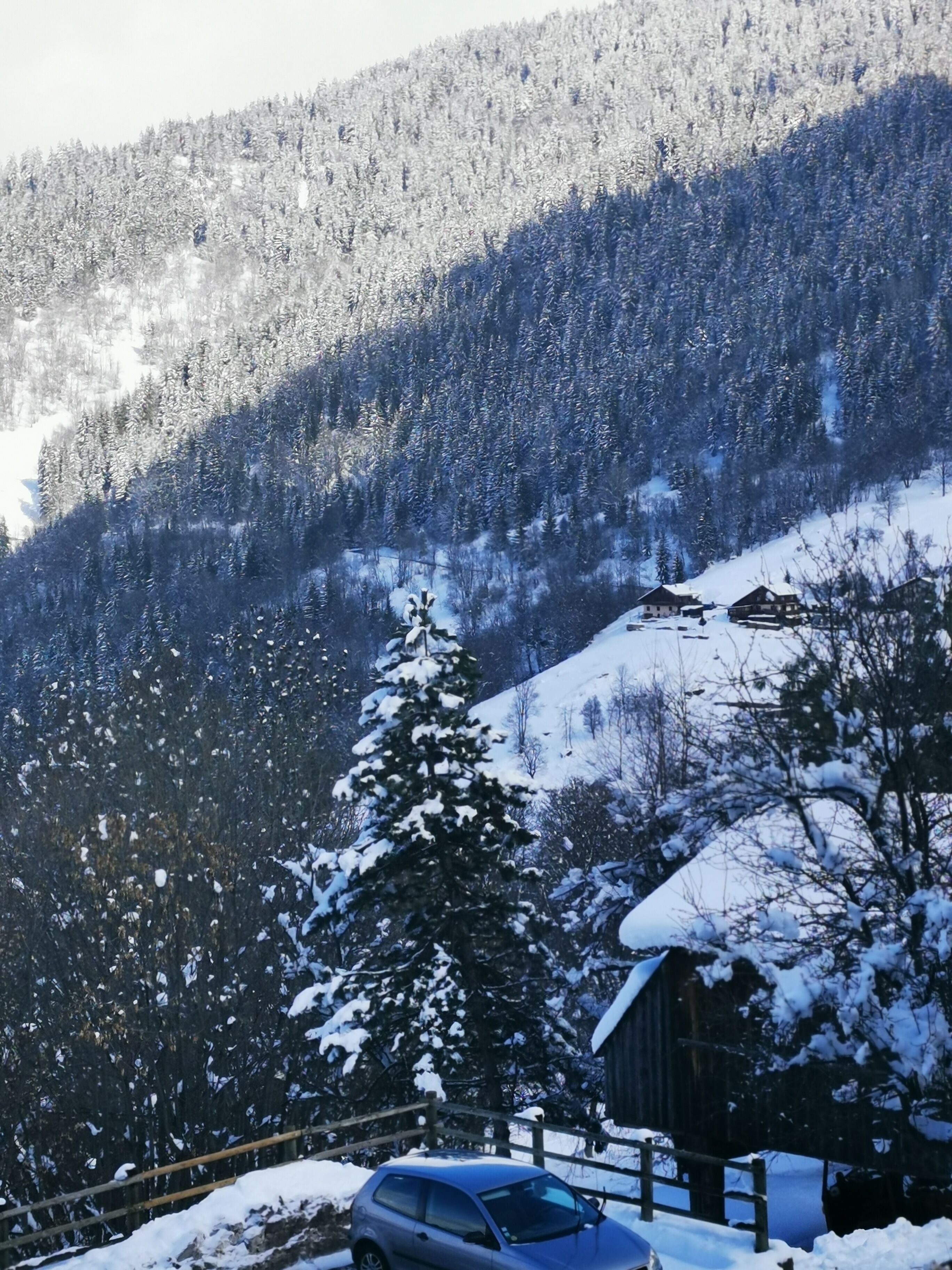 A rustic cabin with a snow-covered roof, surrounded by a wooden fence and a parked car.