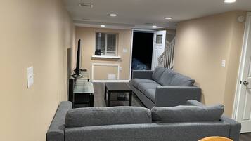Una sala de estar con un sofá gris, una mesita y un televisor. Se ve una escalera que conduce a otra habitación.