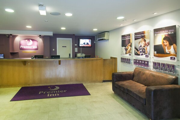 A hotel reception area with a wooden reception desk, a television mounted on the wall, and a couch. - Premier Inn Leeds City Centre - Wellington Street (Leeds)