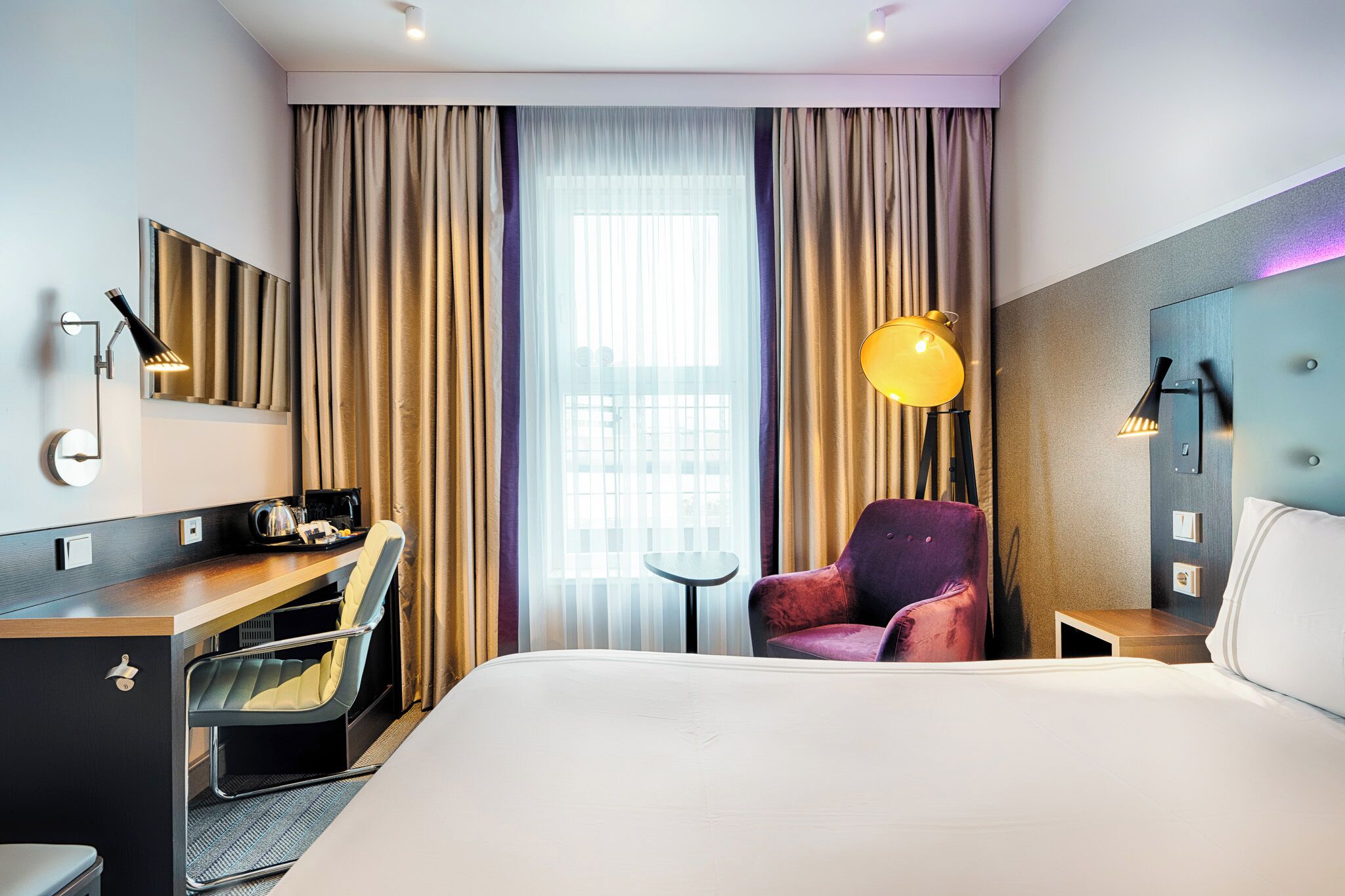 A hotel room with a bed, a desk, a chair, a small table, a lamp, and a window with curtains.