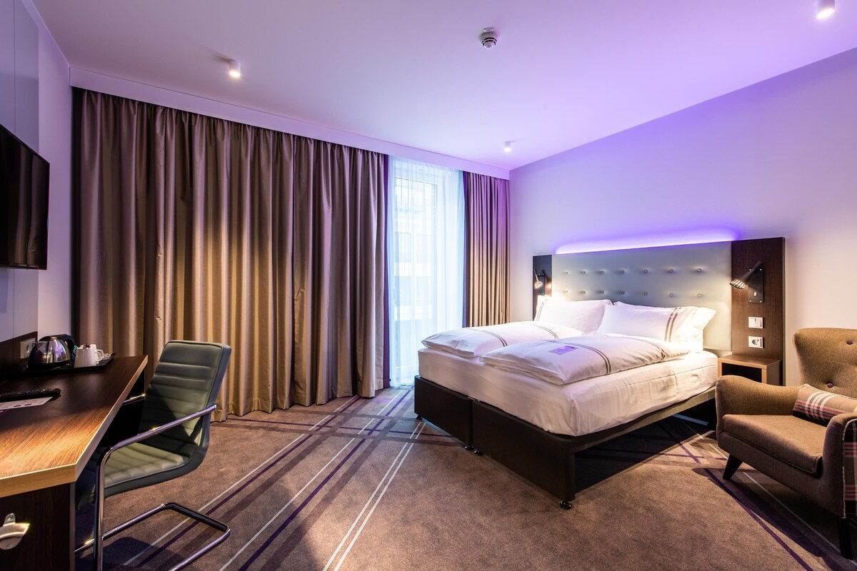Habitación de hotel con cama, escritorio, silla y un ventanal con cortinas.