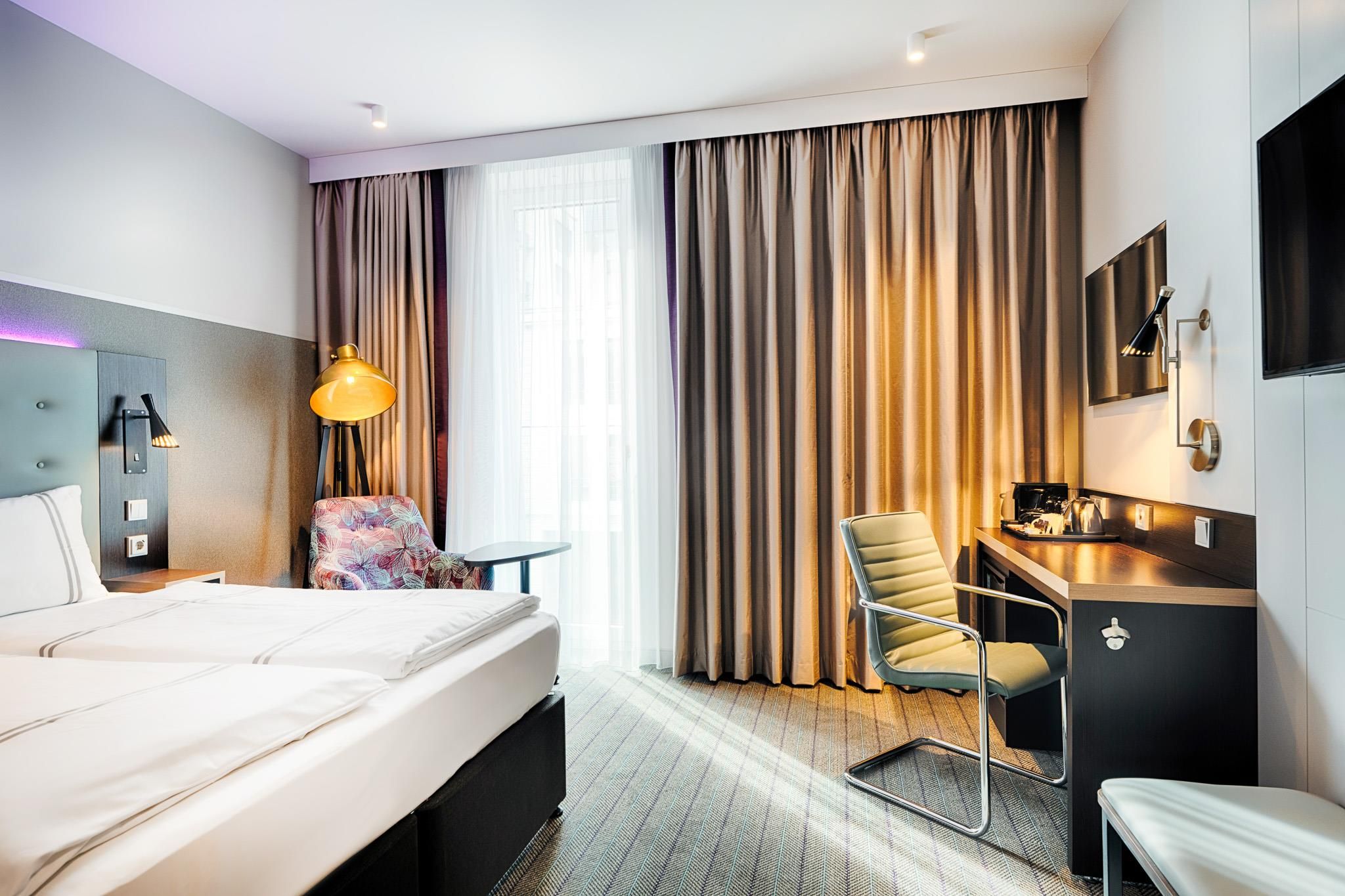 A hotel room with a bed, a desk, a chair, a TV, and a window with curtains.