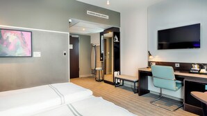 Quarto de hotel com cama, escrivaninha, cadeira, televisão e chuveiro.