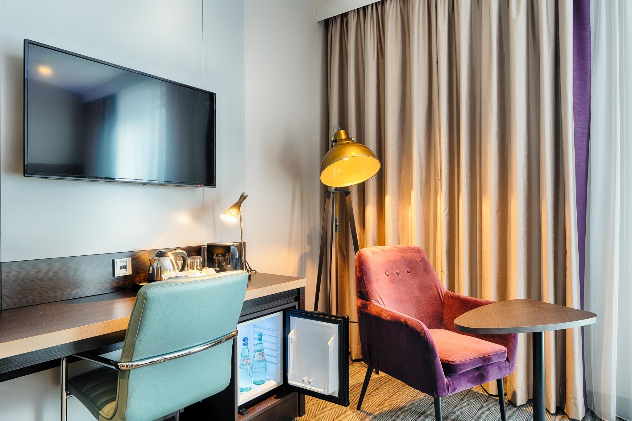 A hotel room with a flat-screen TV, a desk with a lamp, a chair, a small table, and a minibar.