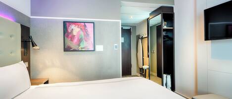 A modern hotel room with a large bed, a painting on the wall, a TV mounted on the wall, and a walk-in closet.