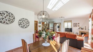 Een eetgedeelte met een ronde tafel, stoelen en een bloevas. Achterin staat een bank en een televisie.