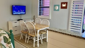 En matplats med ett träbord och vita stolar, en tv som visar ett nyhetsprogram och ett fönster med persienner.