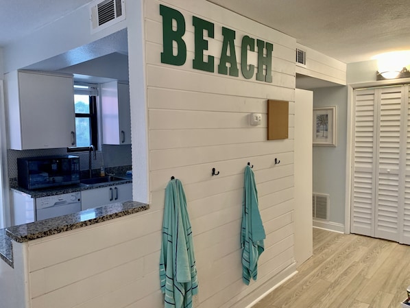 A kitchen with a microwave, sink, and cabinets. There is a wall-mounted towel rack with towels. A sign on the wall reads "BEACH".
