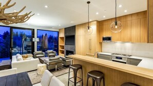 A modern kitchen with wooden cabinets, a central island, bar stools, and a large window overlooking a forested area. - $7 Million Aspen 4BR Residence - Brand New Construction (Aspen)
