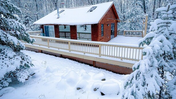 Eine Holzhütte mit einer Holzterrasse und einem mit Schnee bedeckten Dach, umgeben von schneegefüllten Bäumen.